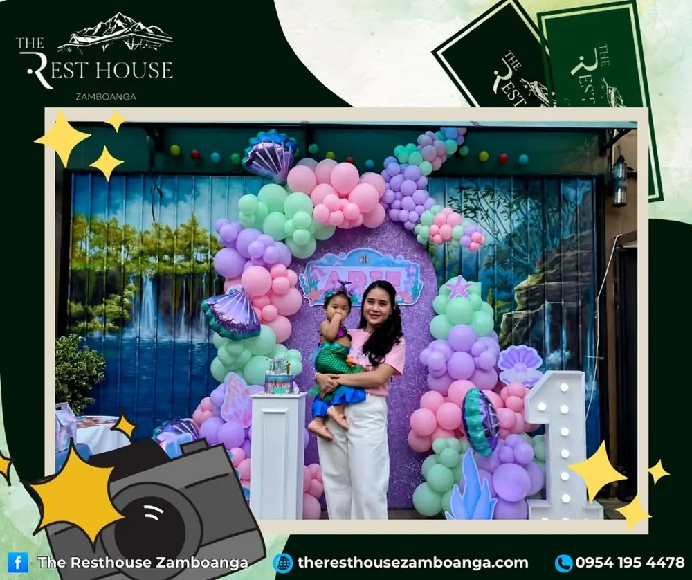 A mother holding her daughter in a mermaid costume, in front of a pastel balloon arch for a first birthday